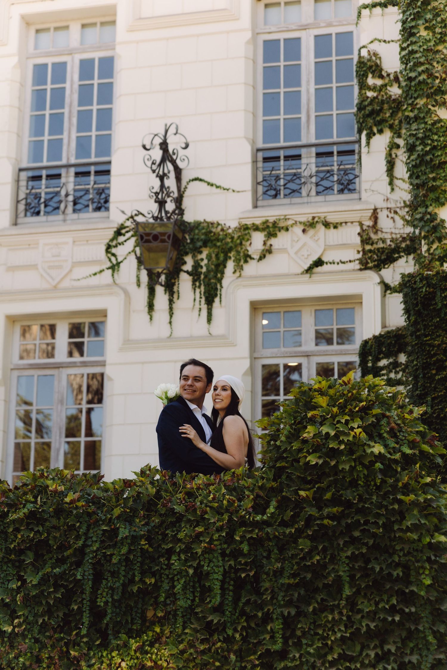 Bride and groom posing together against ivy-draped white building facade with ornate ironwork.