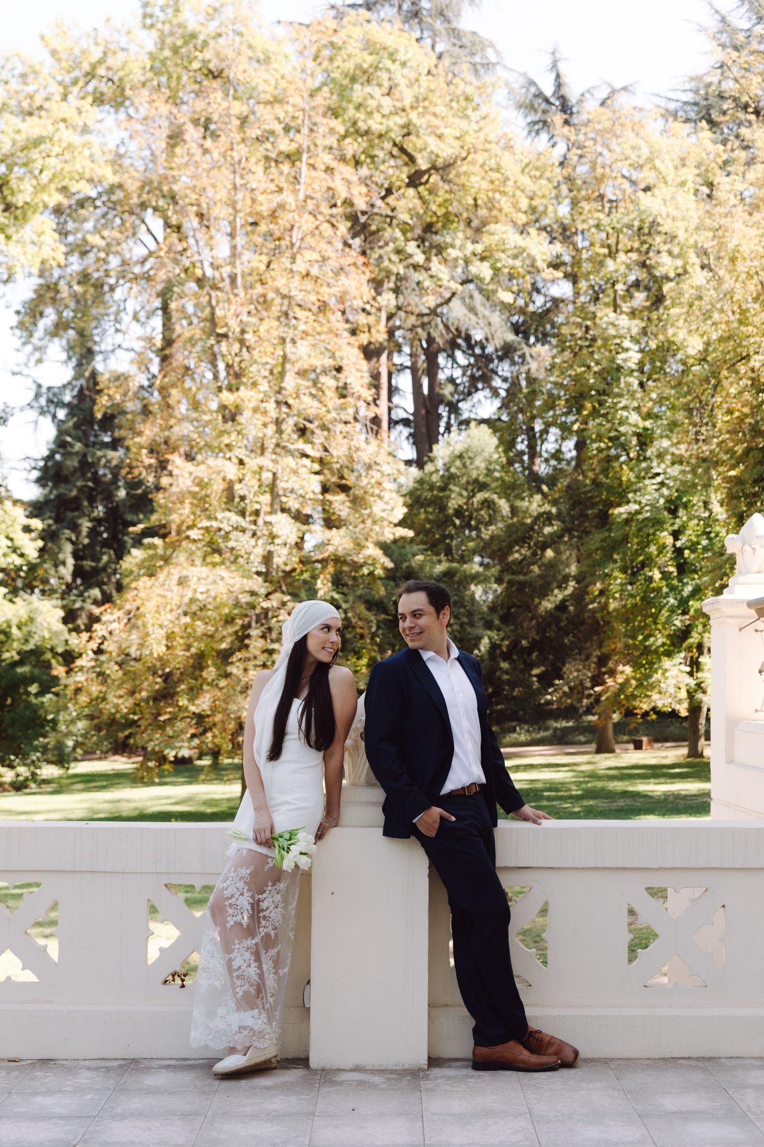 Bride in lace dress and groom leaning against white balustrade with lush green trees behind.