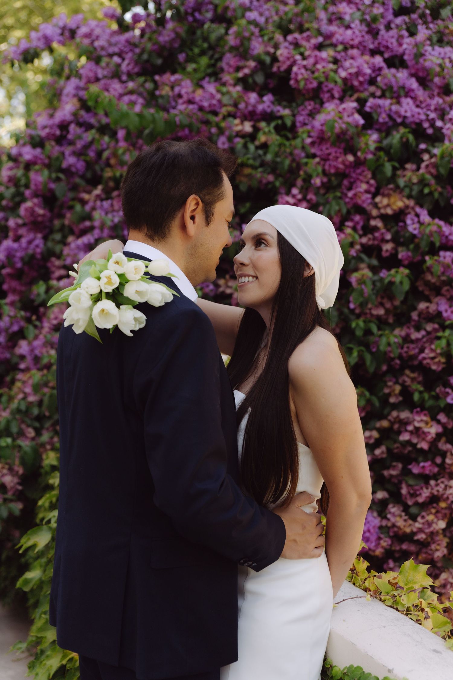 Smiling bride in white dress and head wrap embracing groom before vibrant purple flower wall.