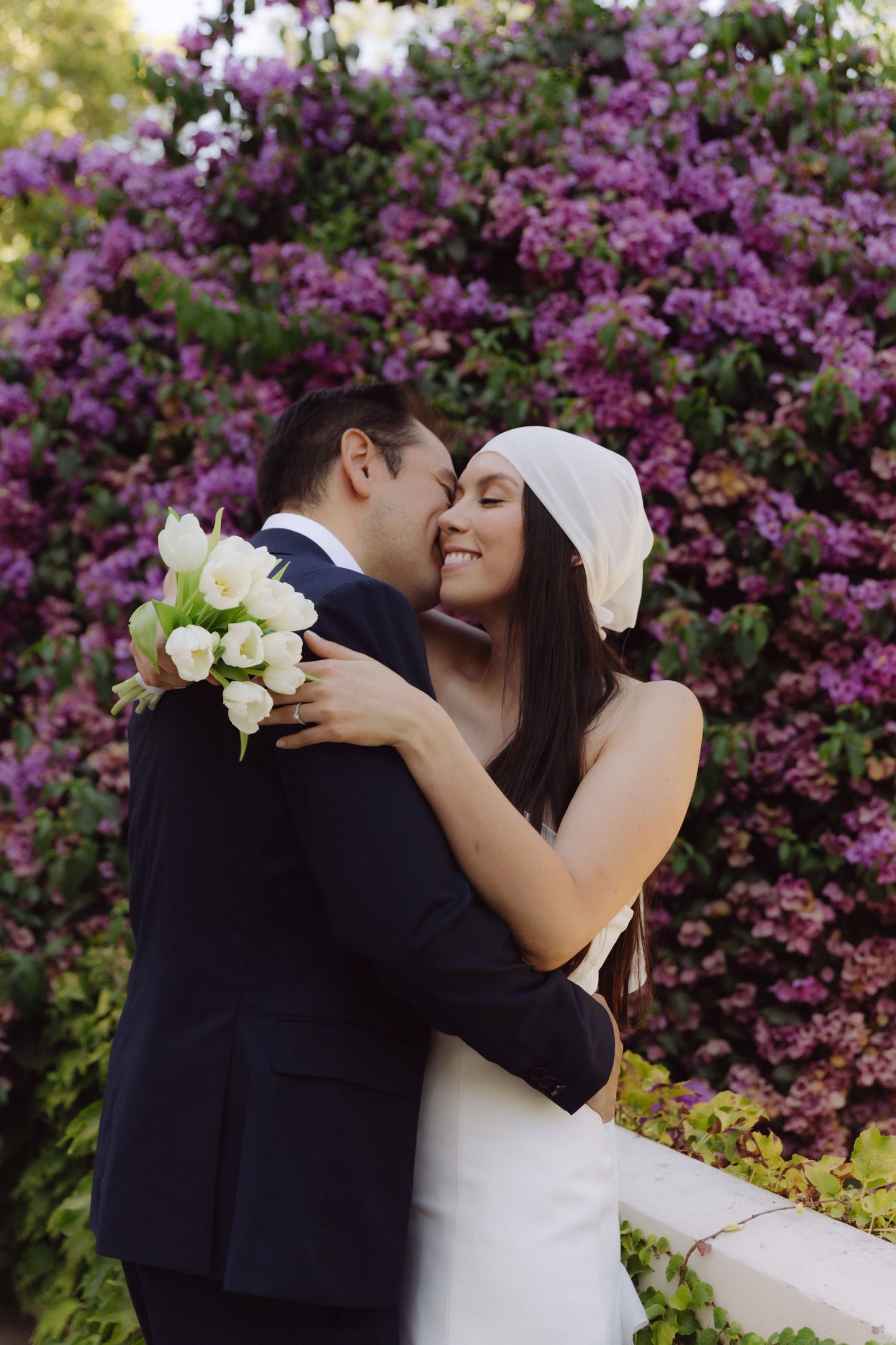 Groom kissing smiling bride holding white tulip bouquet against blooming purple flower backdrop.