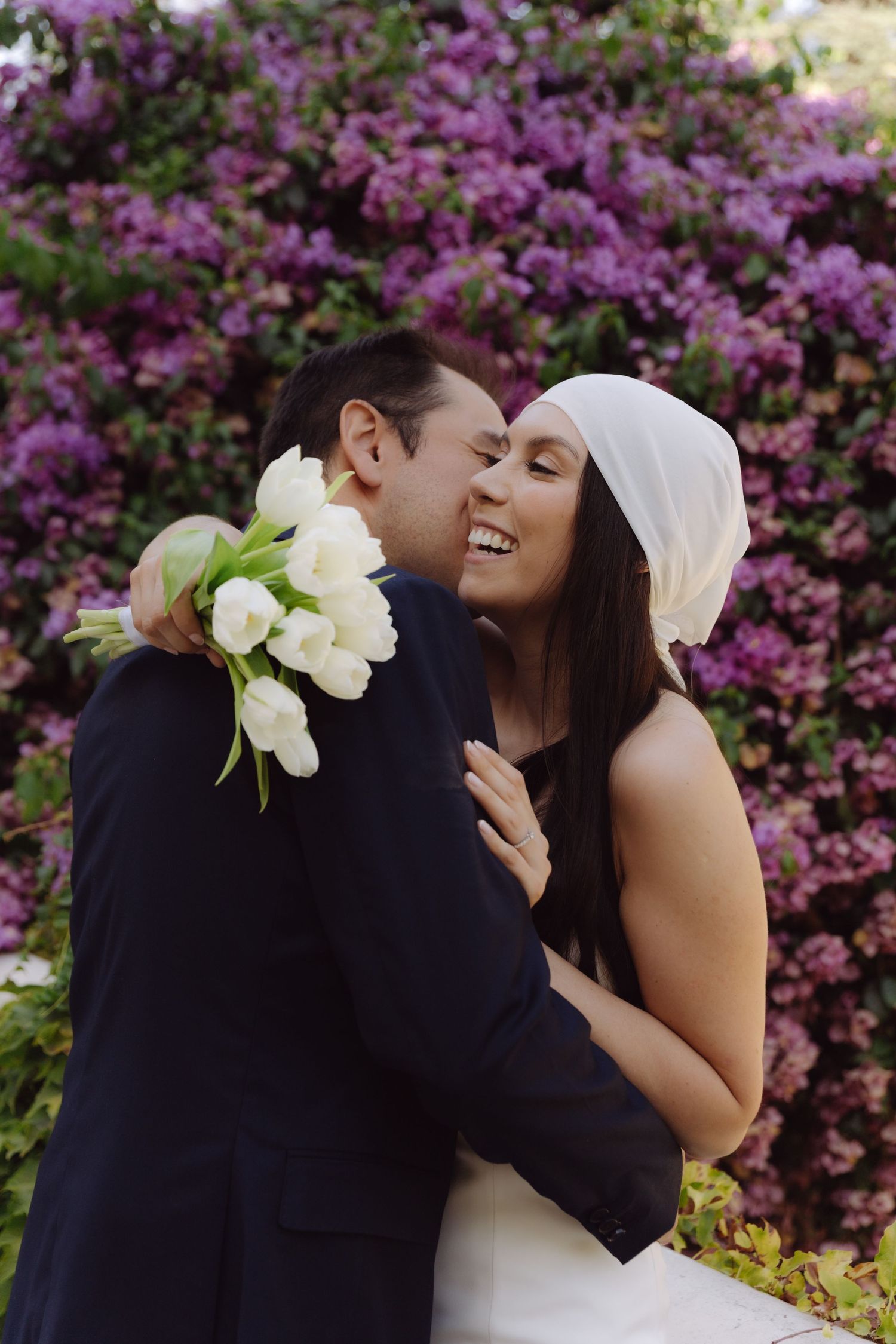 Laughing bride holding white tulips embracing groom before a stunning purple flowering hedge.