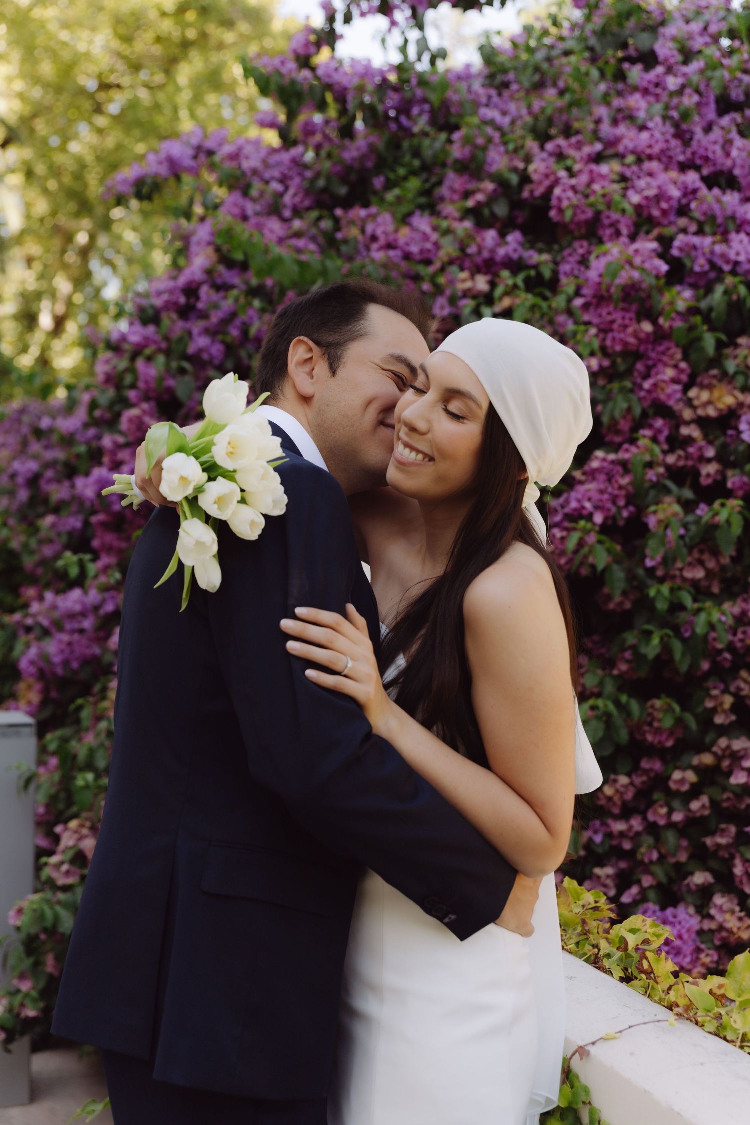 Couple sharing tender embrace with white tulip bouquet before vibrant purple blossom backdrop.