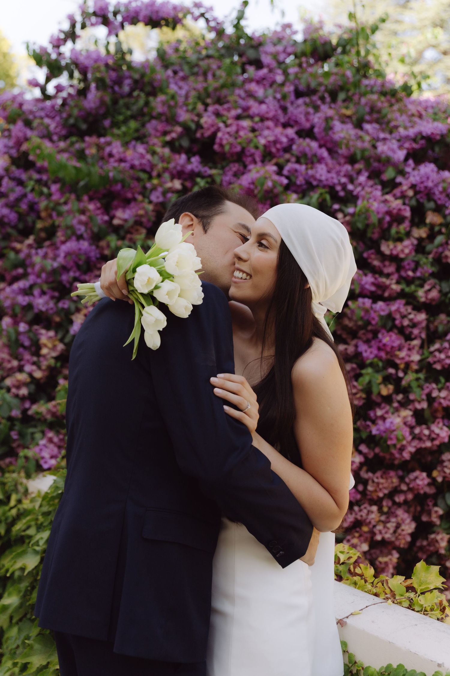 Bride and groom embrace before purple flowering bushes, bride holding white tulip bouquet wearing white headband.