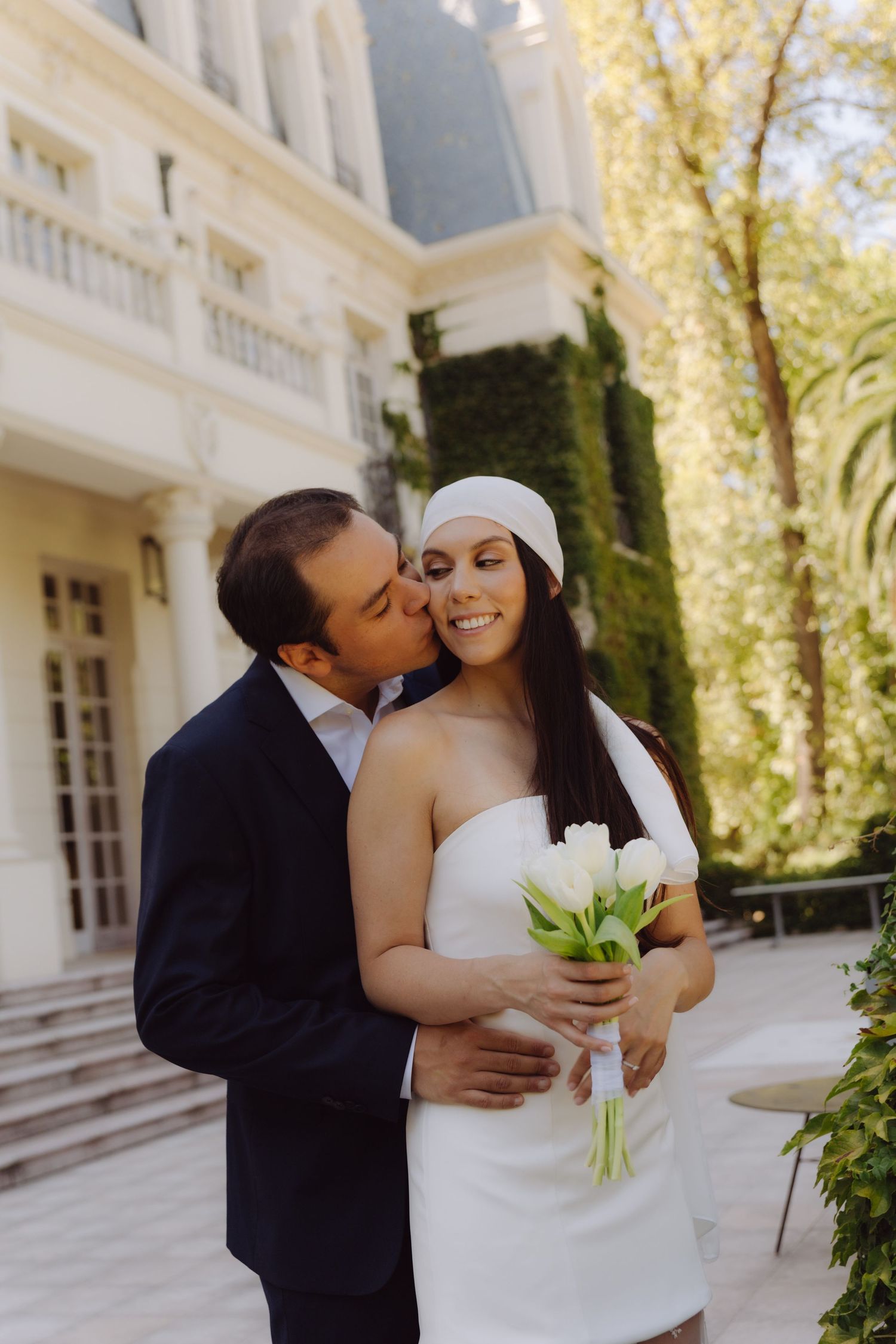 Groom in navy suit kisses bride's cheek as she smiles holding white tulips outside elegant mansion.