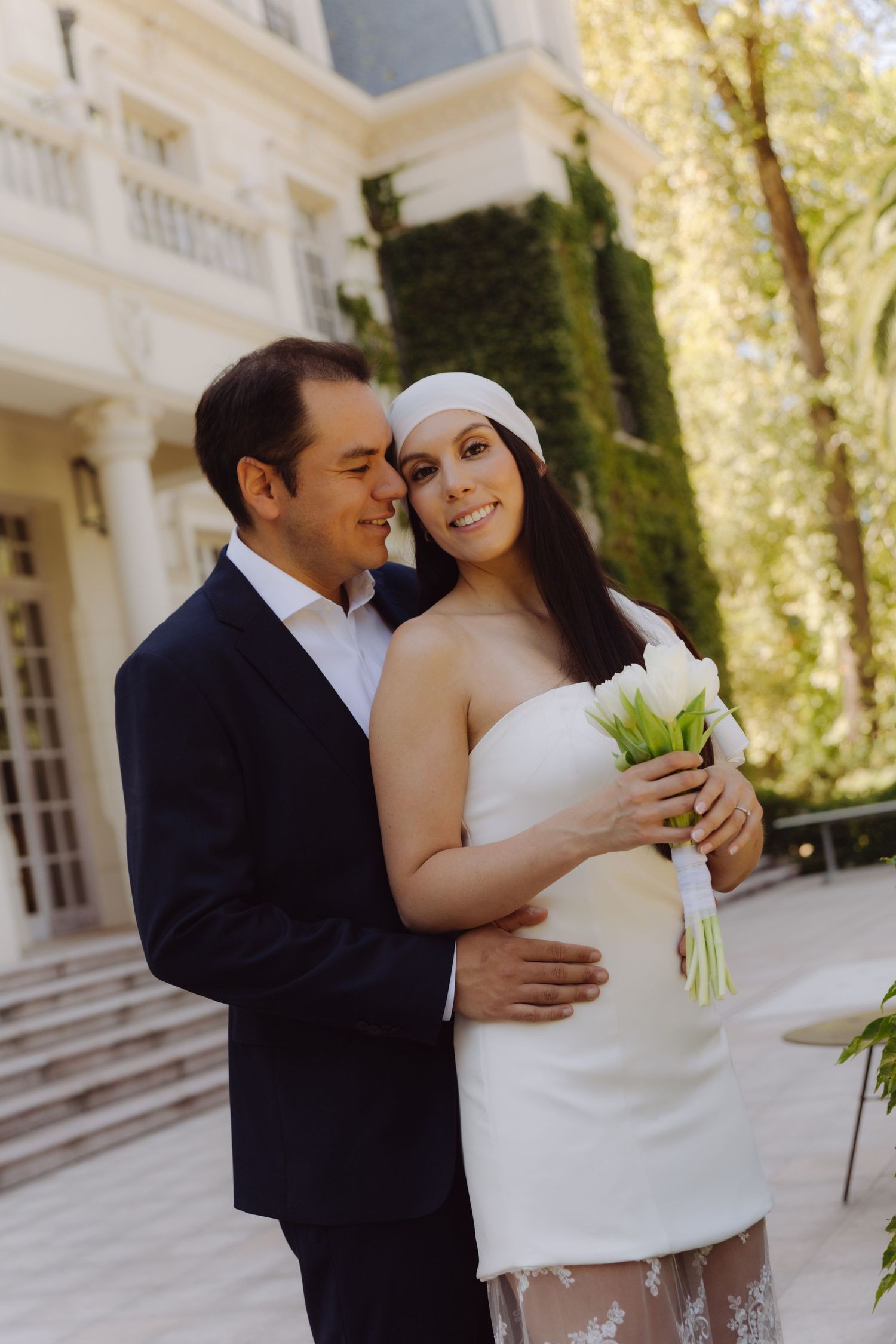 Smiling bride in white strapless dress and headband embraced by groom holding white tulip bouquet outdoors.