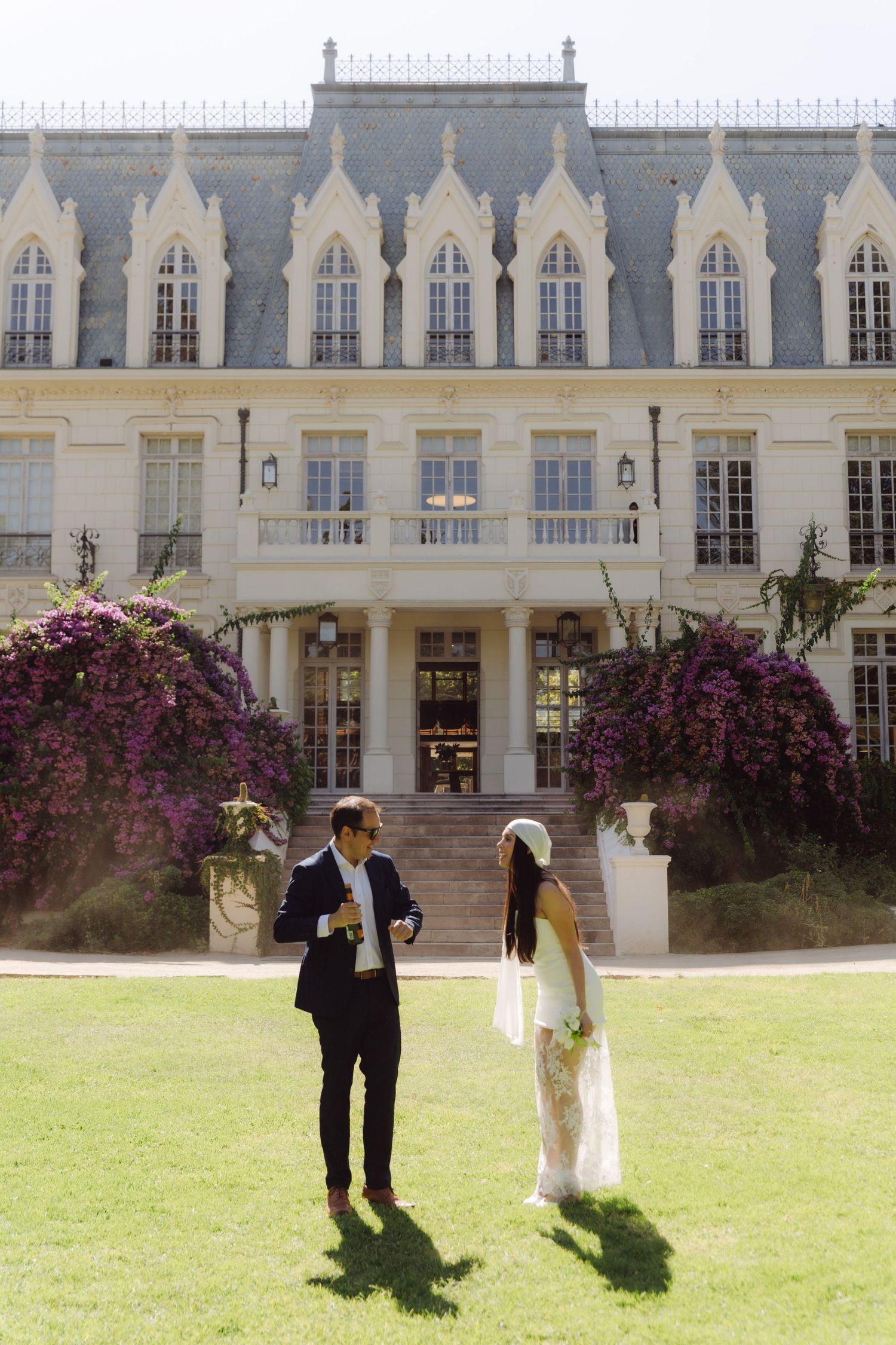 Bride and groom stand on green lawn before grand Gothic-style mansion adorned with purple bougainvillea.