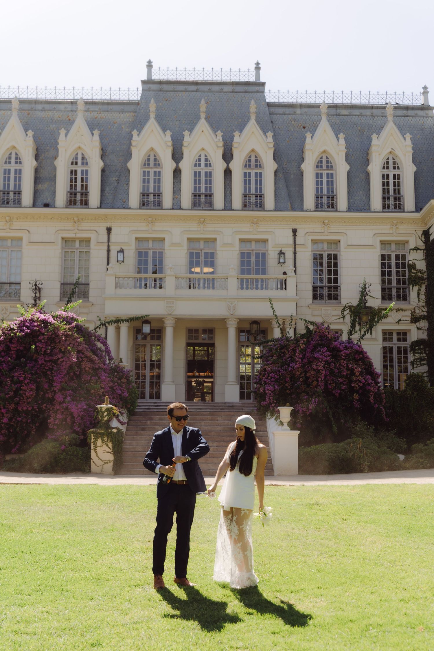 Newlyweds holding hands on sunlit lawn in front of elegant chateau with purple flowering vines.