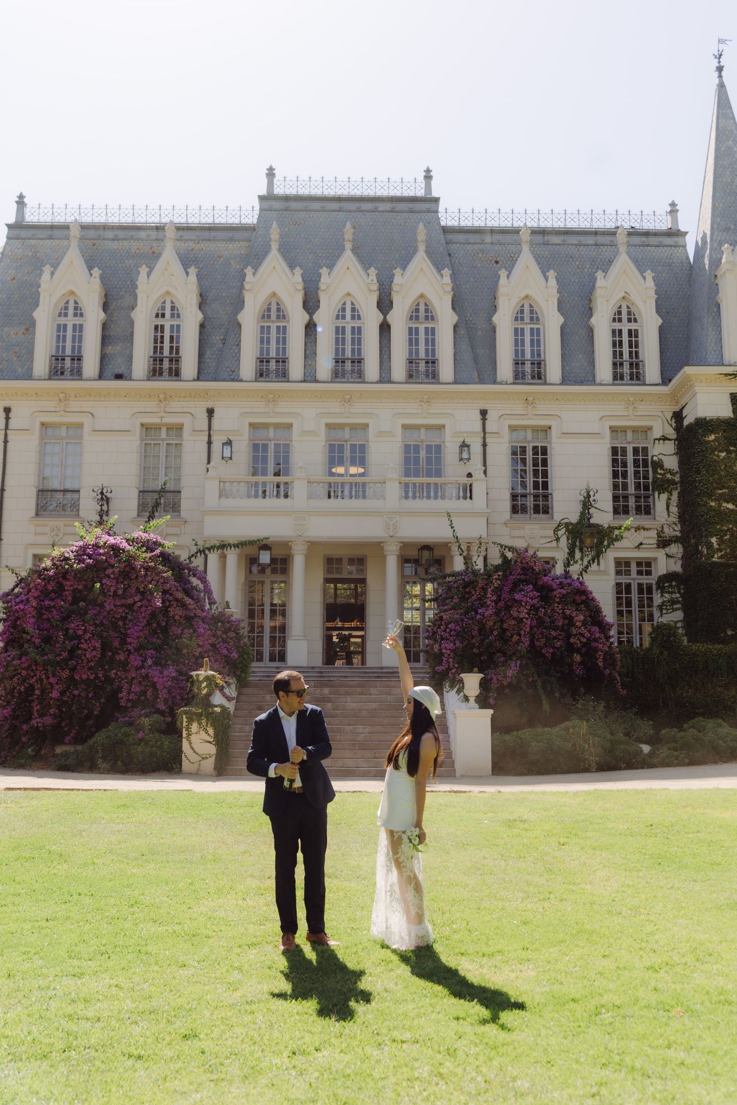 Bride raises bouquet joyfully while groom checks phone on lawn before ornate French-style chateau.