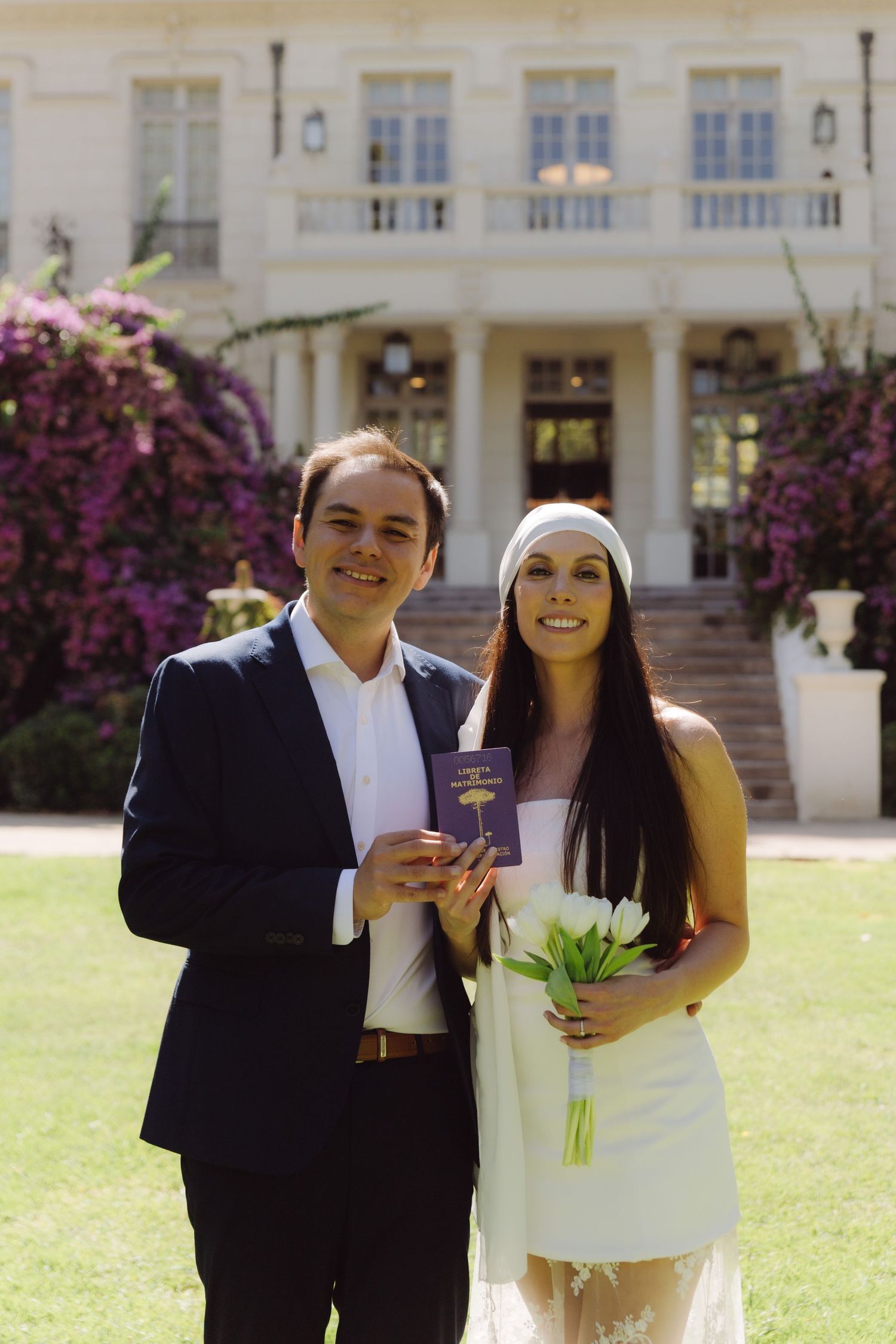 Newlyweds proudly display purple marriage certificate and white tulip bouquet before bougainvillea-adorned mansion.