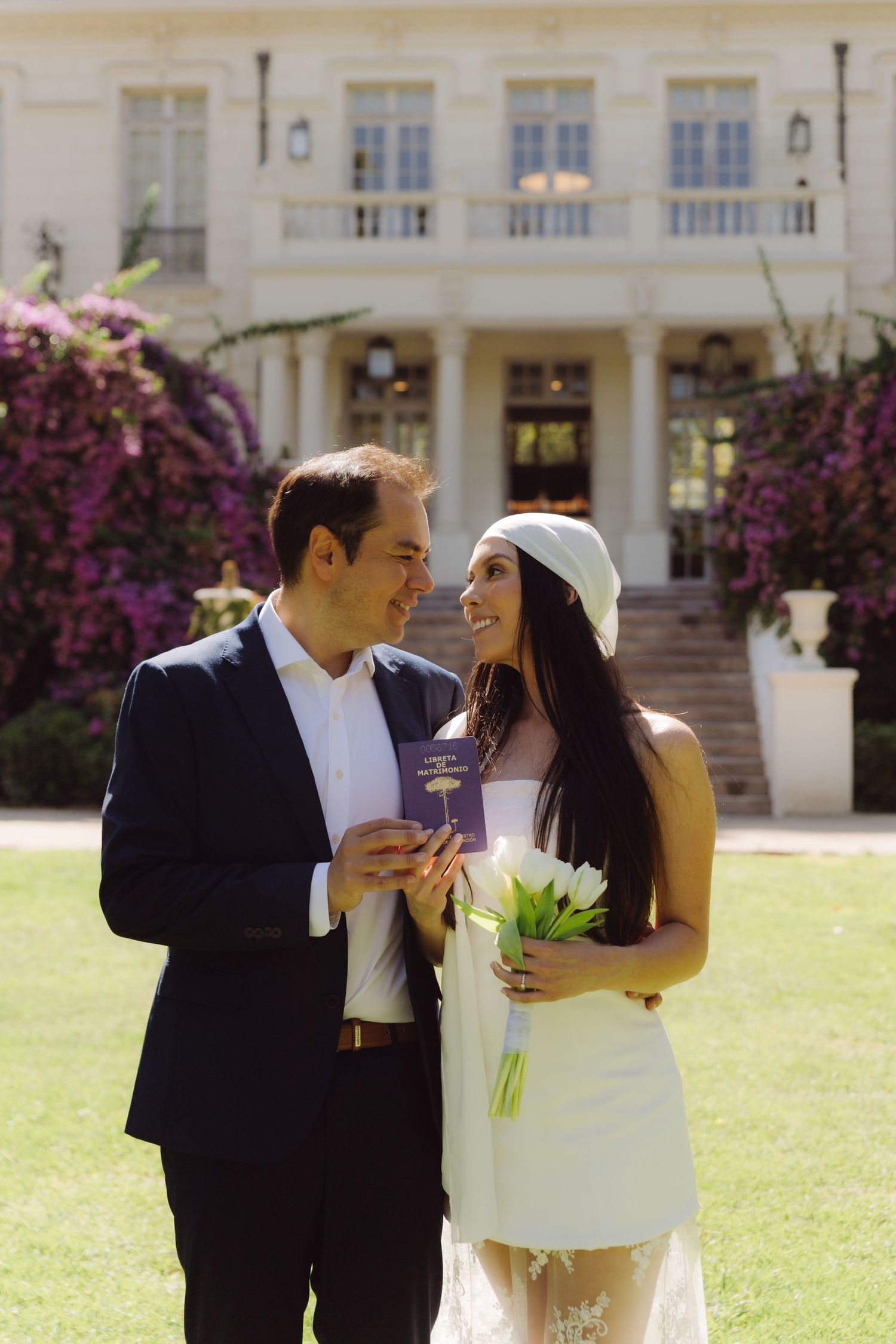 Smiling couple holds purple marriage booklet and white tulips on lawn outside elegant floral estate.