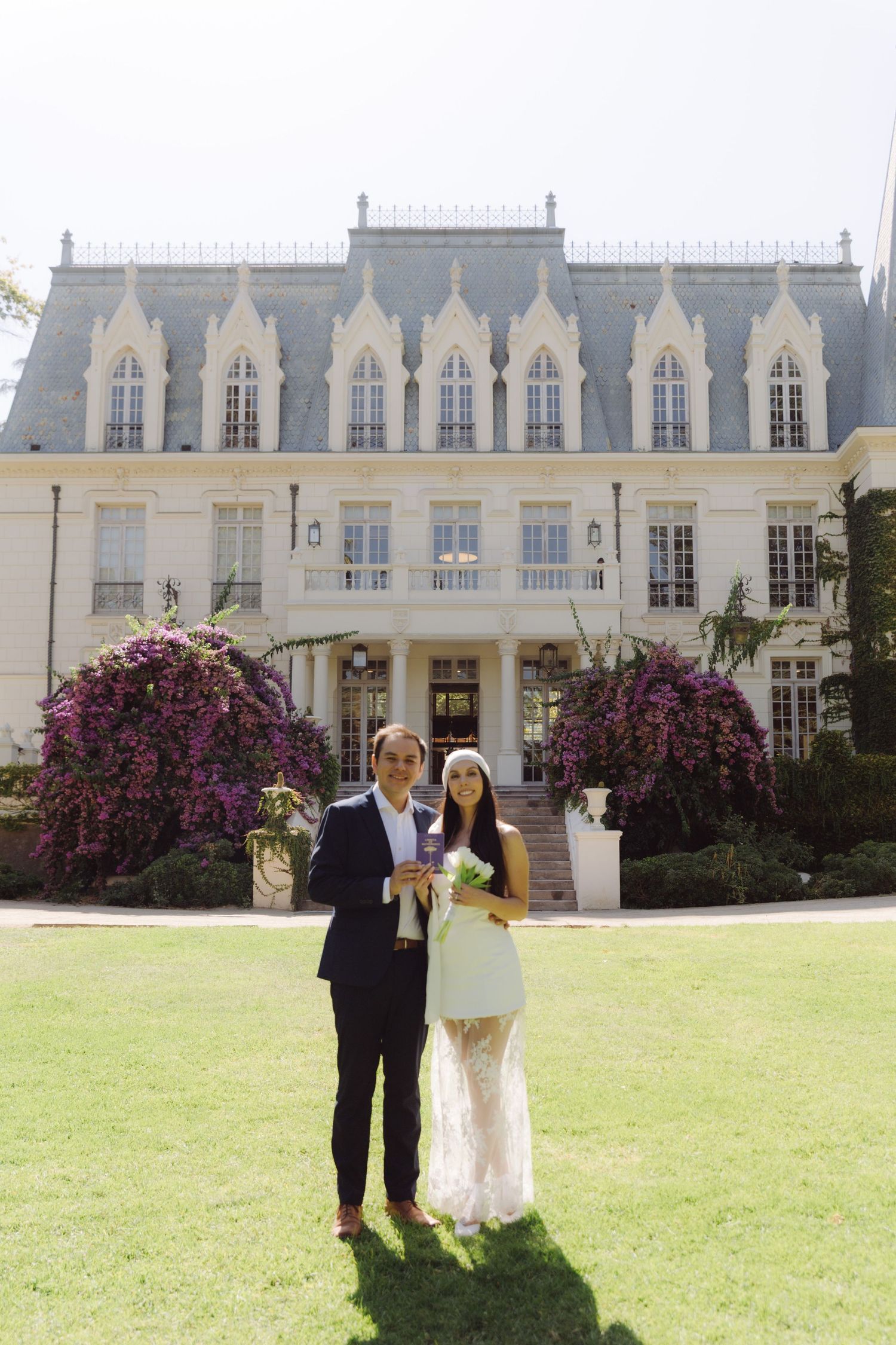 Bride and groom pose together on sunny lawn before grand French chateau with purple bougainvillea.