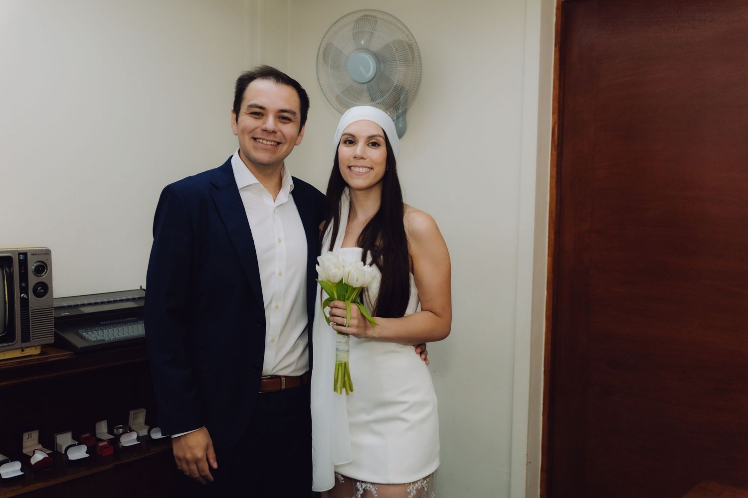 Newlyweds pose indoors after civil ceremony, bride holding white tulip bouquet wearing white headband dress.
