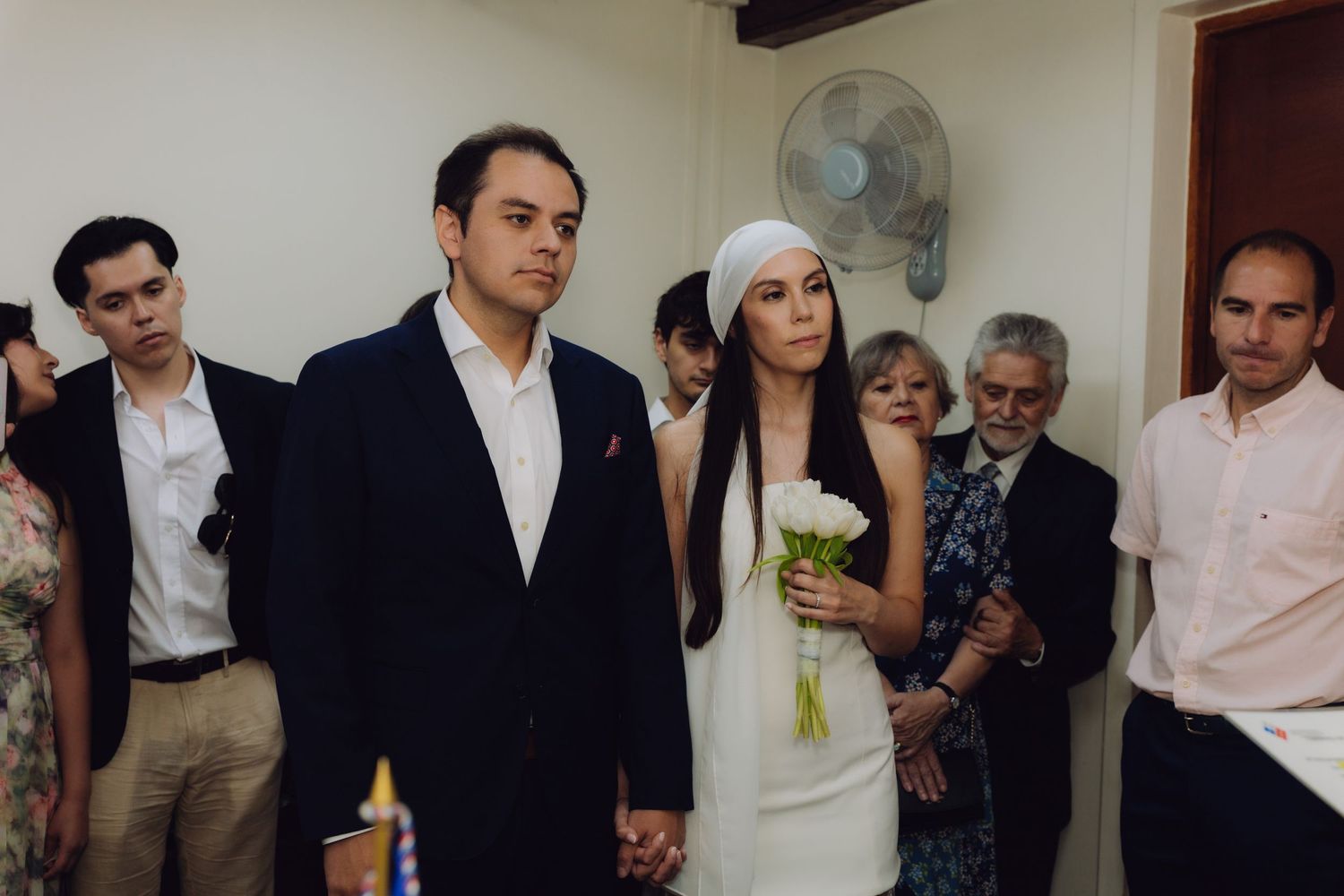 Couple holding hands at intimate wedding ceremony, bride in white dress and headpiece holding bouquet, guests watching.