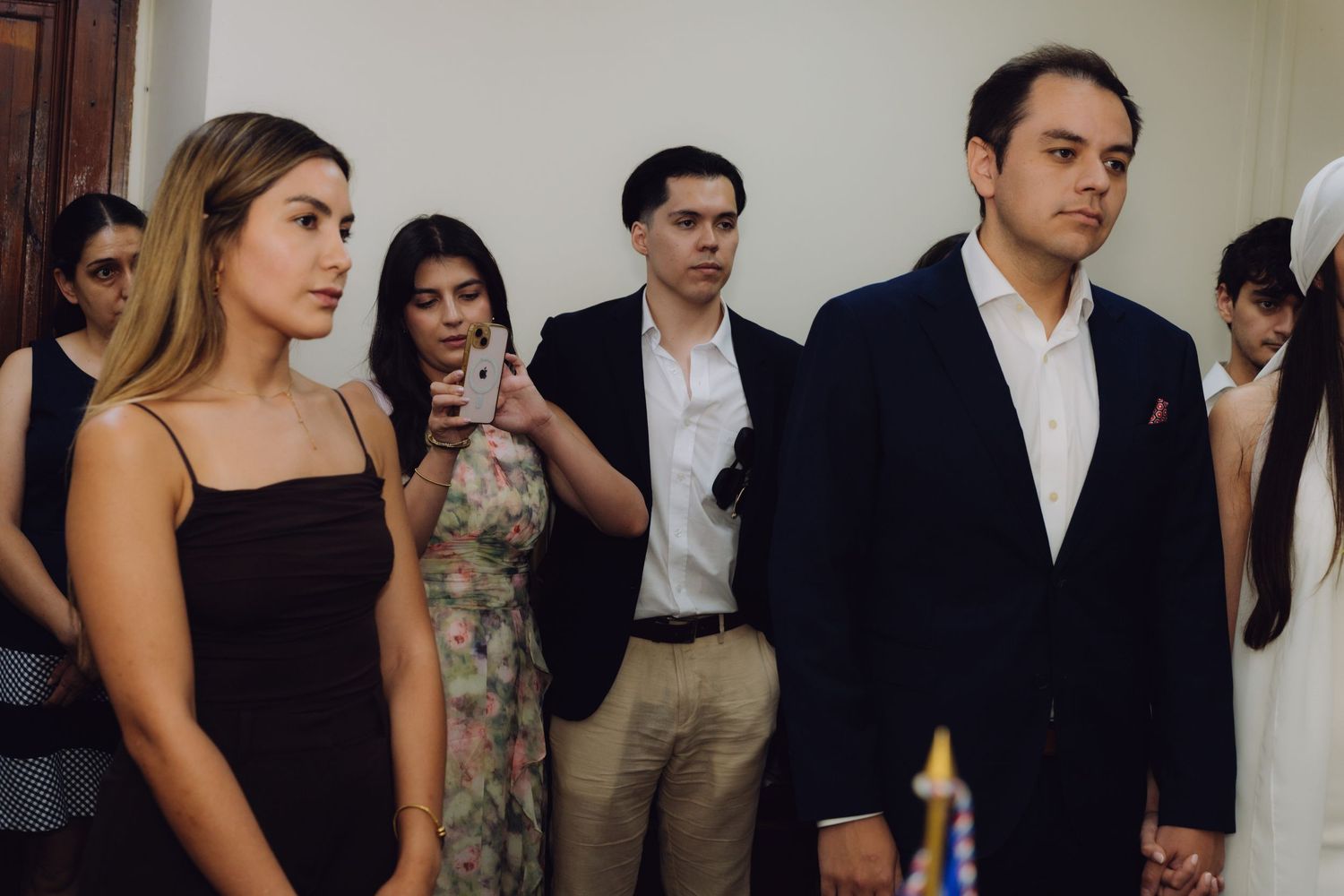 Elegantly dressed wedding guests standing in formal venue, woman in black dress and man in dark suit in foreground.