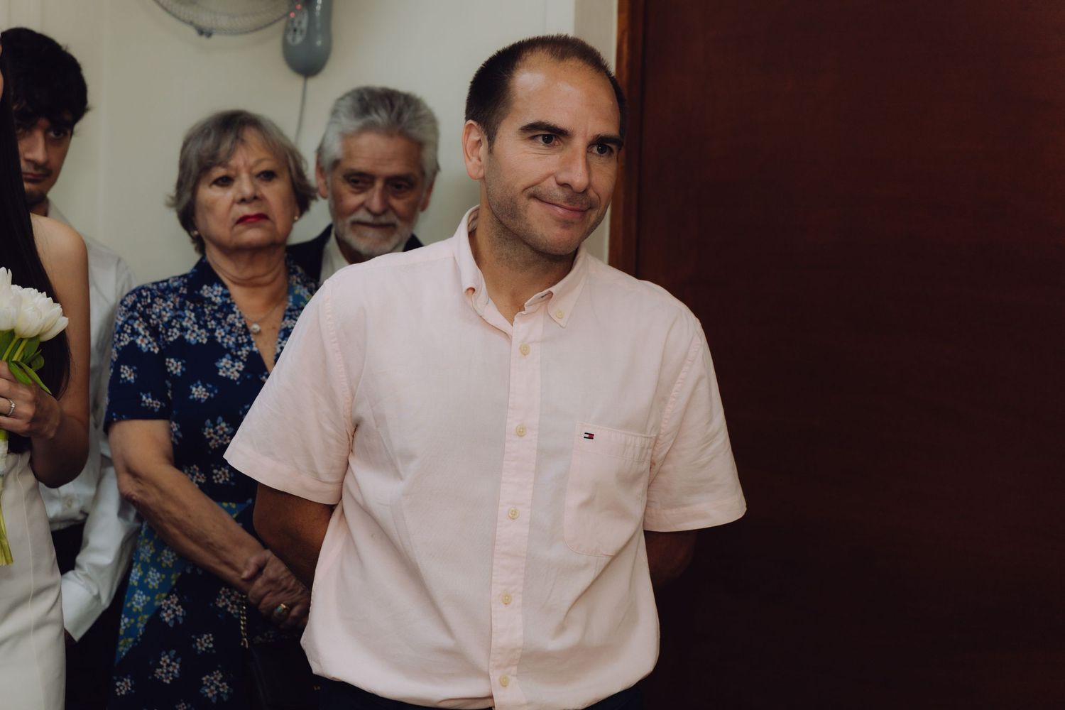 Smiling man in pink shirt at wedding celebration with elderly couple visible in background.