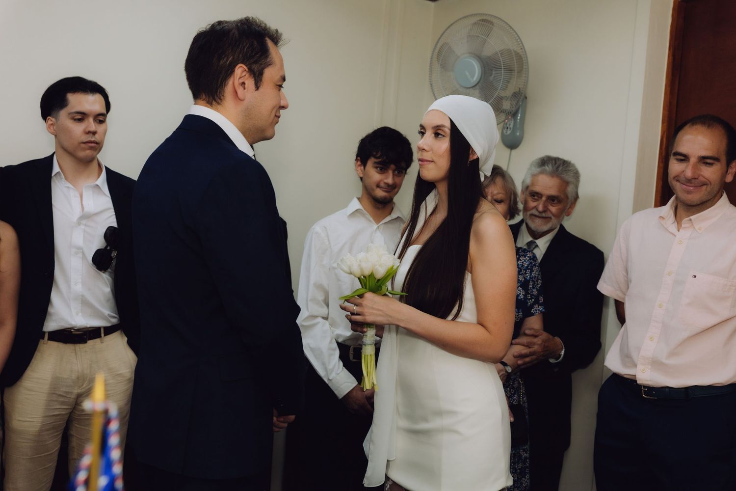 Groom in navy suit exchanging vows with bride holding white bouquet, surrounded by wedding guests indoors.