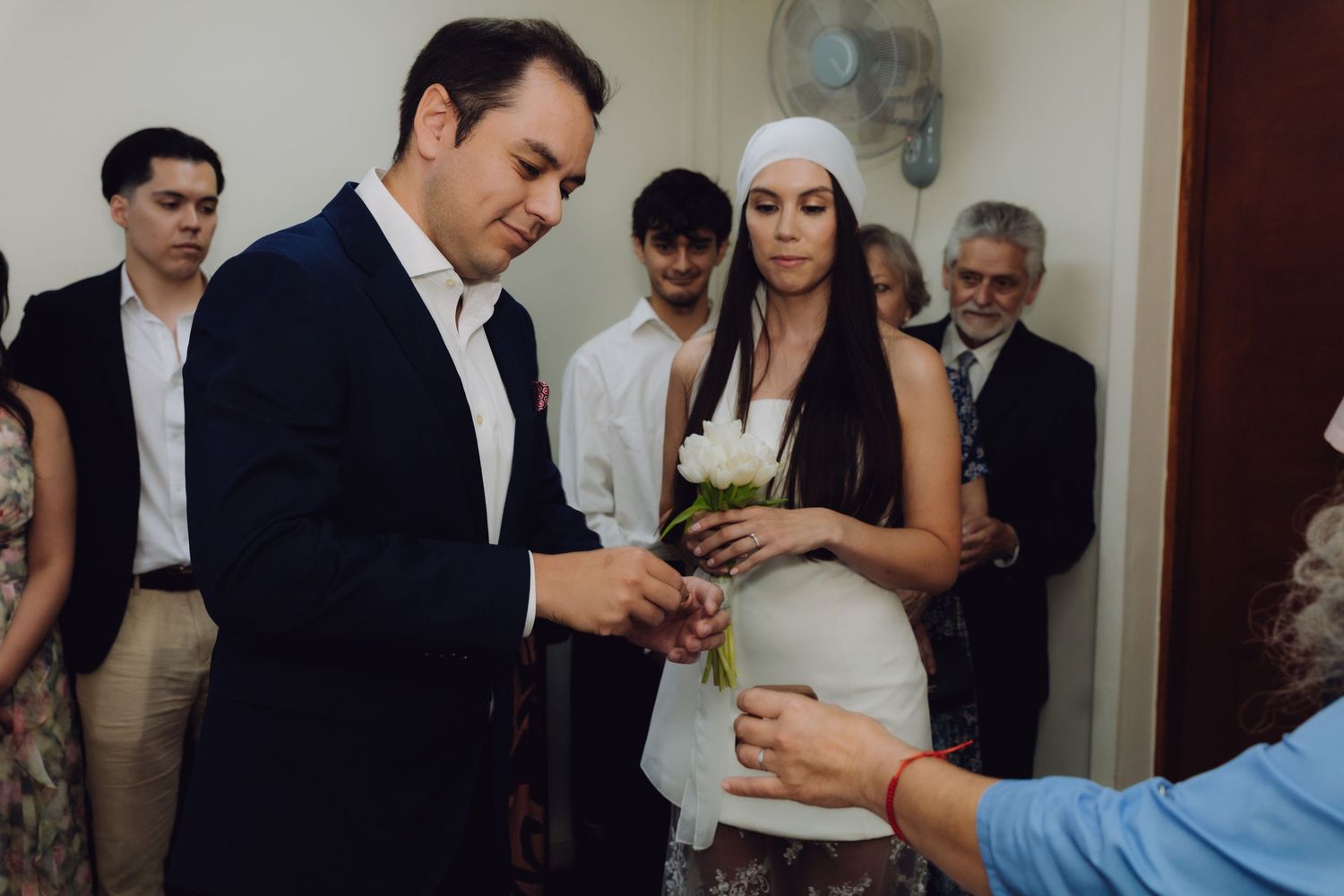 Groom in navy suit receiving wedding ring from officiant, bride in white dress and headpiece standing nearby.