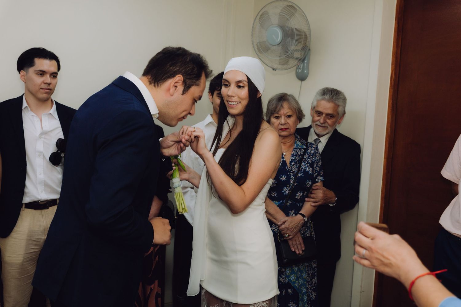 Groom placing wedding ring on brides finger during intimate indoor ceremony with family guests watching.