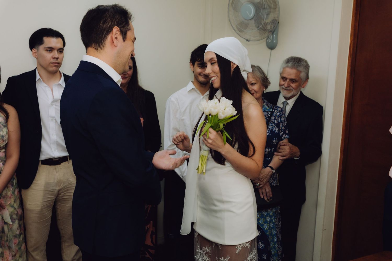 Groom sliding wedding ring onto brides finger as guests smile during intimate indoor wedding ceremony.