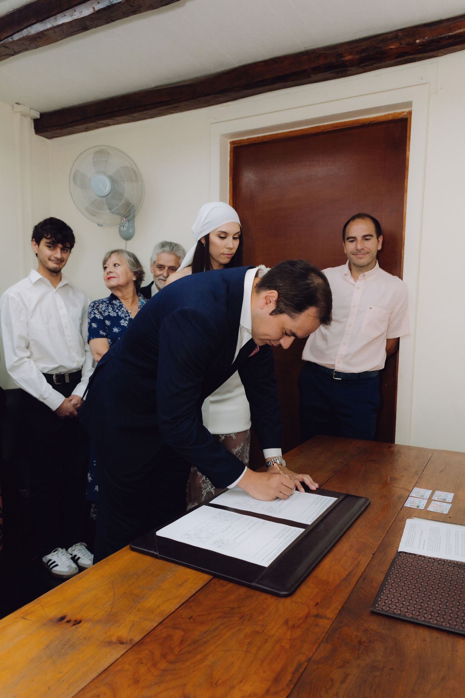 Groom in navy suit signing official wedding documents at wooden table, bride and guests watching ceremony.