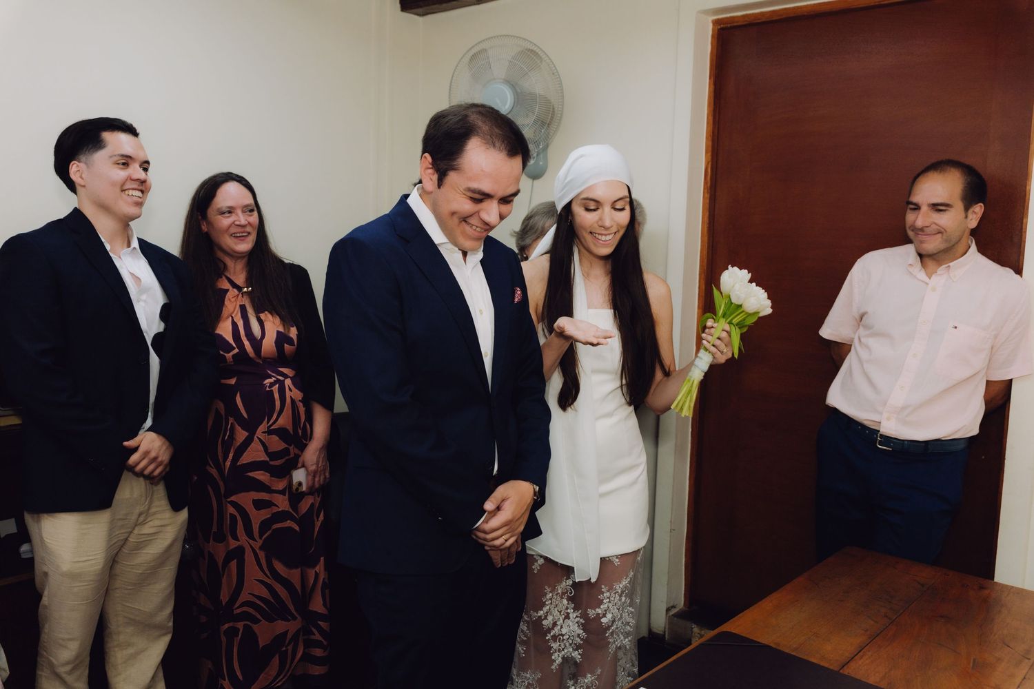 Bride in white dress and headpiece stands with groom and family at civil wedding.