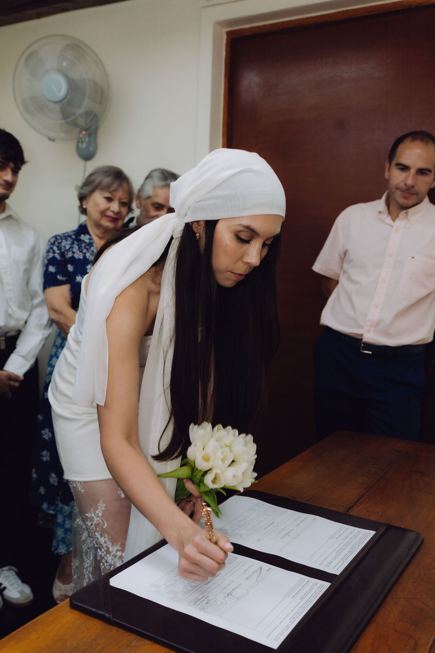 Bride in white dress and headpiece signs wedding documents at a wooden desk.