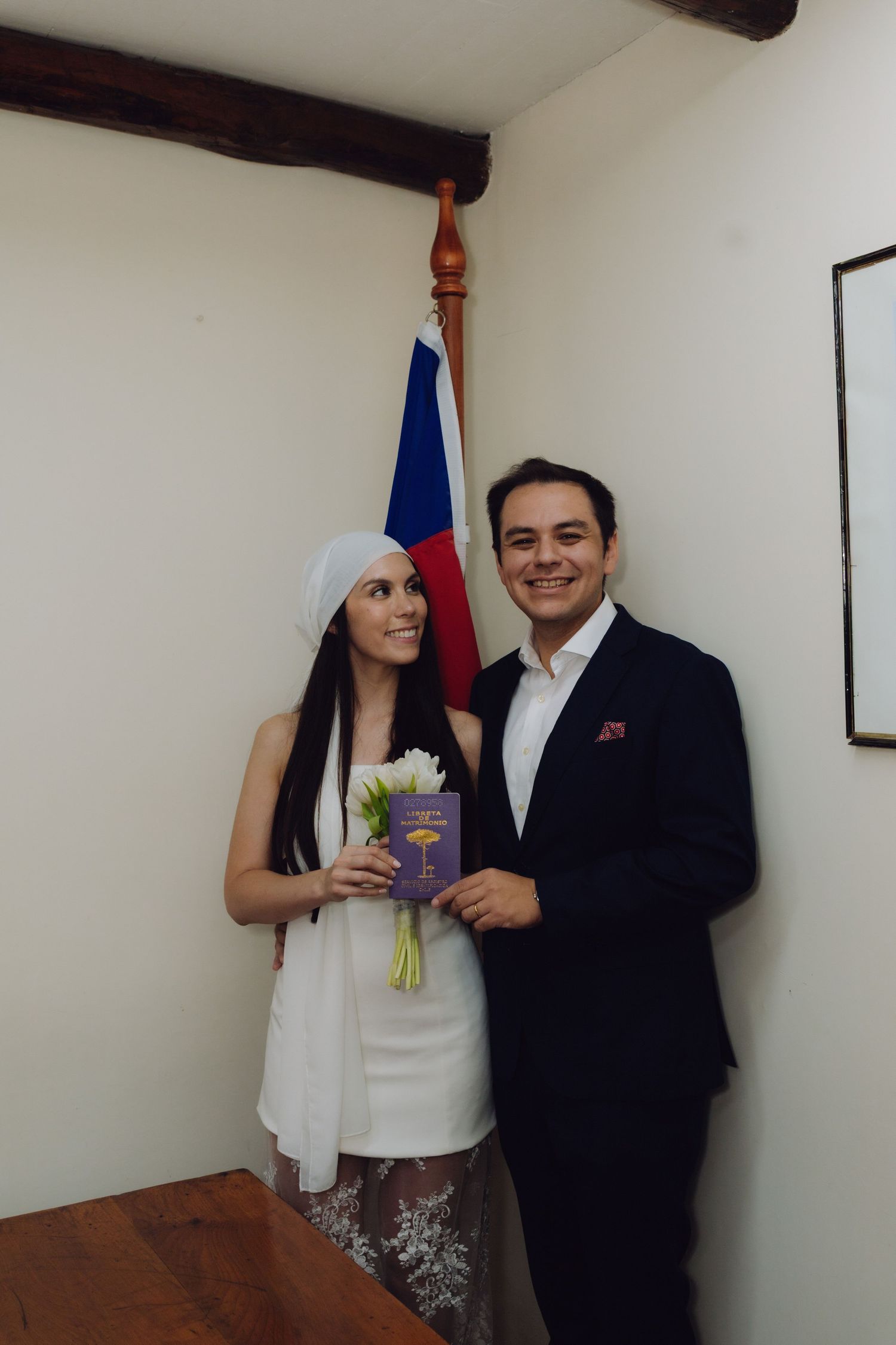 Bride in white dress and headpiece holds tulips beside smiling groom in navy suit near a flag.