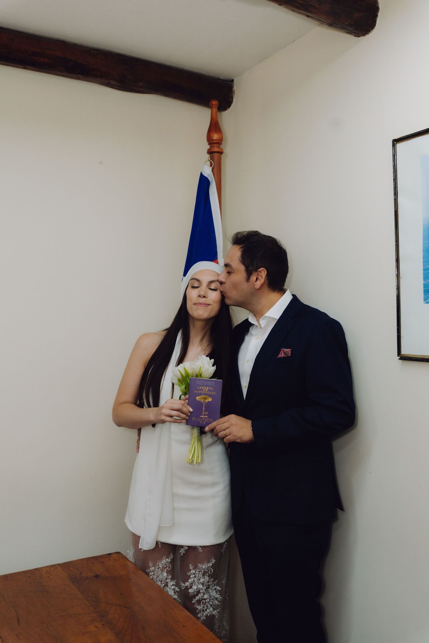 Groom in navy suit kisses bride in white dress and headpiece holding white tulips near a flag.