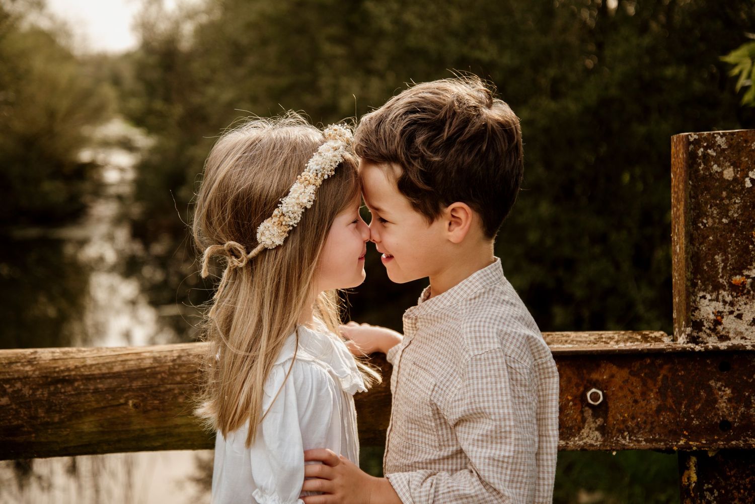 Two young children touching foreheads tenderly near a wooden fence outdoors, girl wearing floral crown in soft natural light.