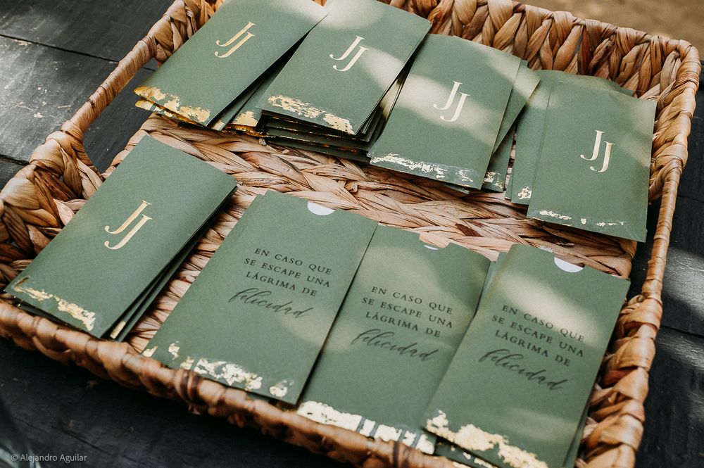 Green wedding programs with gold floral accents and monogram JJ displayed in a wicker basket.