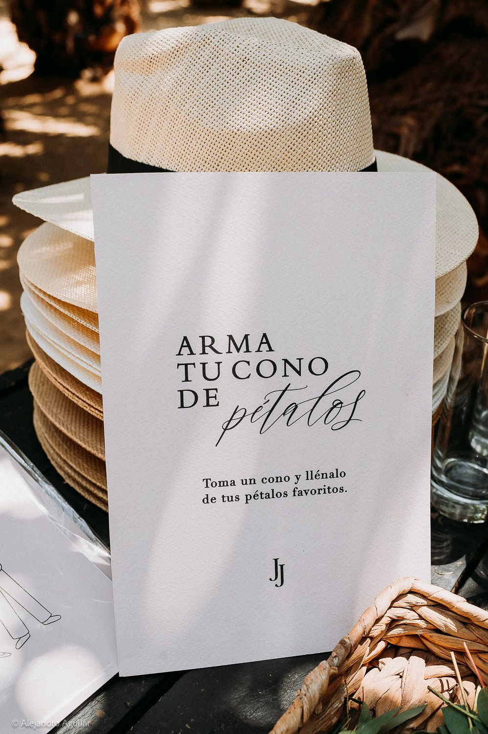 Spanish wedding petal cone sign reading 'Arma Tu Cono de Pétalos' displayed outdoors with straw hat and plates.