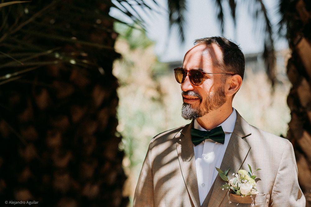 Groom in beige suit, bow tie, sunglasses, and boutonniere stands near palm trees on wedding day.