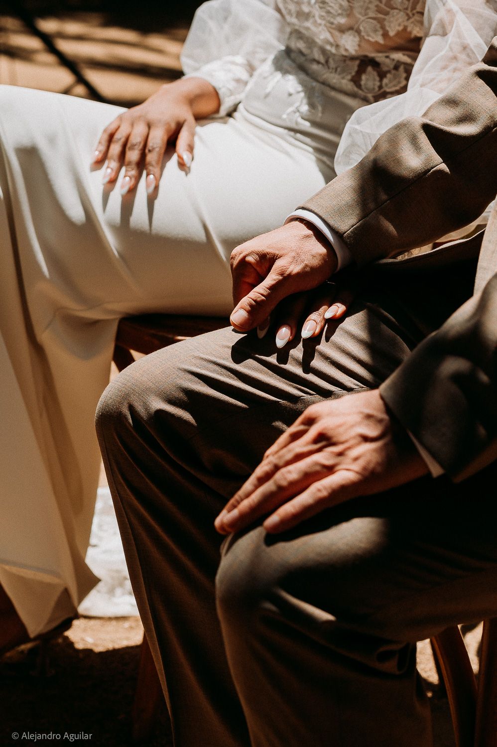 Close-up of bride and groom holding hands while seated, showcasing wedding rings and elegant attire.