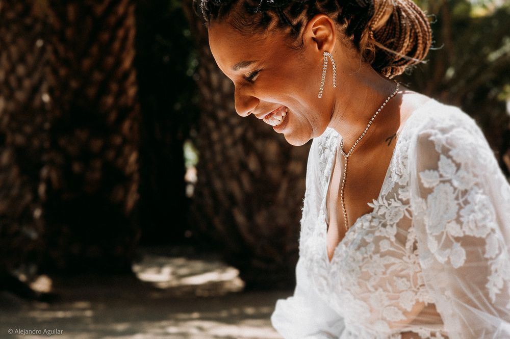 Smiling bride in lace wedding dress with gold earrings and necklace, standing outdoors among trees on her wedding day.