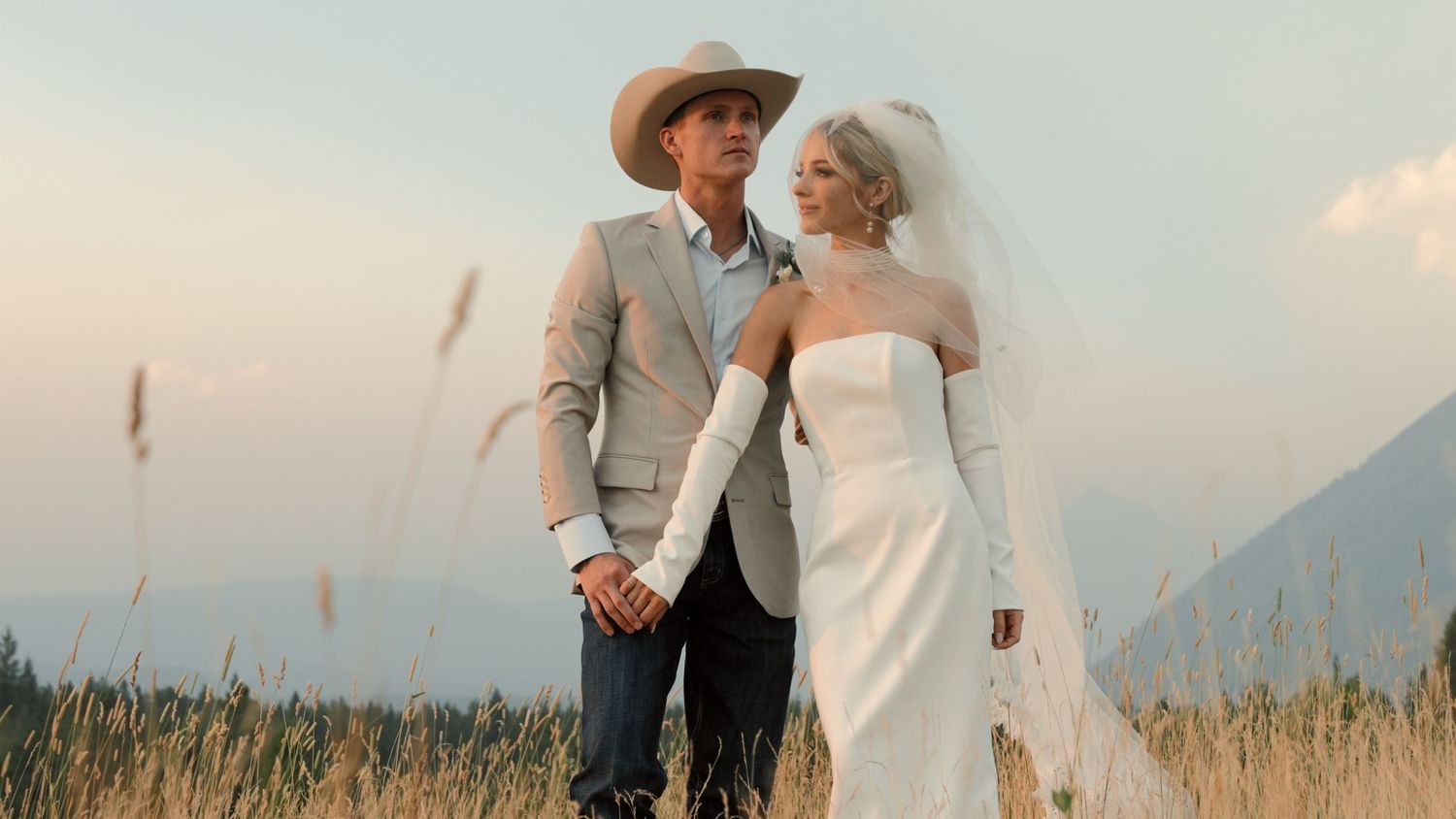 Couple in wedding attire holding hands in a golden field with mountains, bride in white gown and veil, groom in tan suit.
