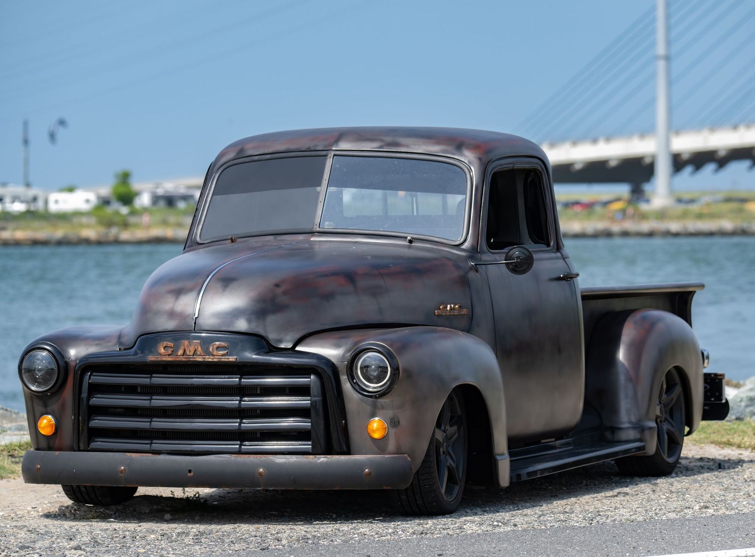 Vintage GMC pickup truck with patina finish parked near waterfront with bridge in background.