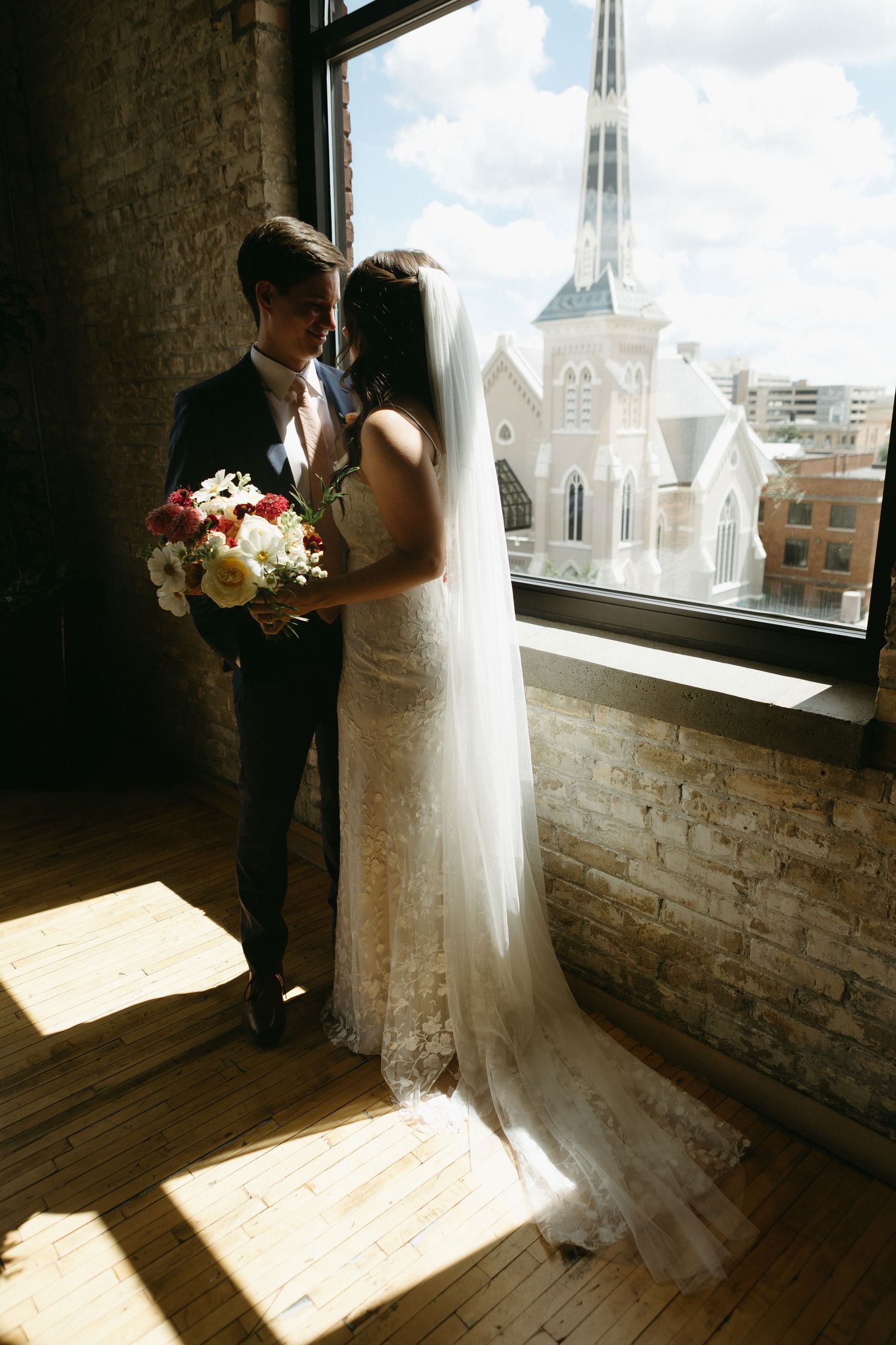 Bride and groom share romantic moment by sunlit window with church view, bride in lace gown with veil holding bouquet.