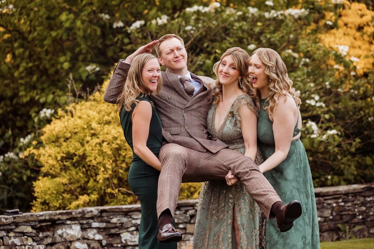 Three women joyfully lift a smiling man in a brown suit outdoors at a wedding celebration.