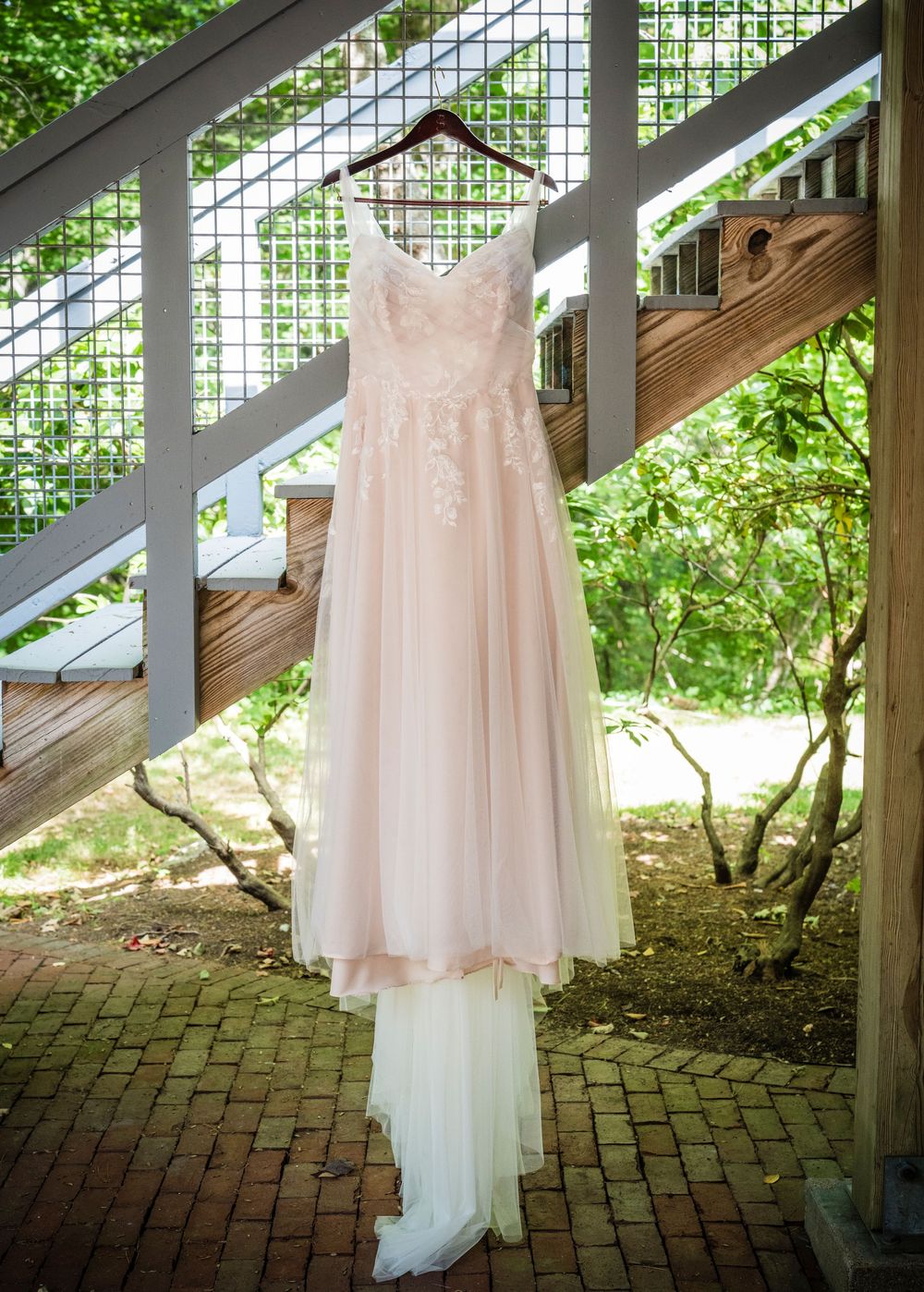Blush pink lace wedding gown hanging on wooden staircase railing outdoors surrounded by lush green trees.
