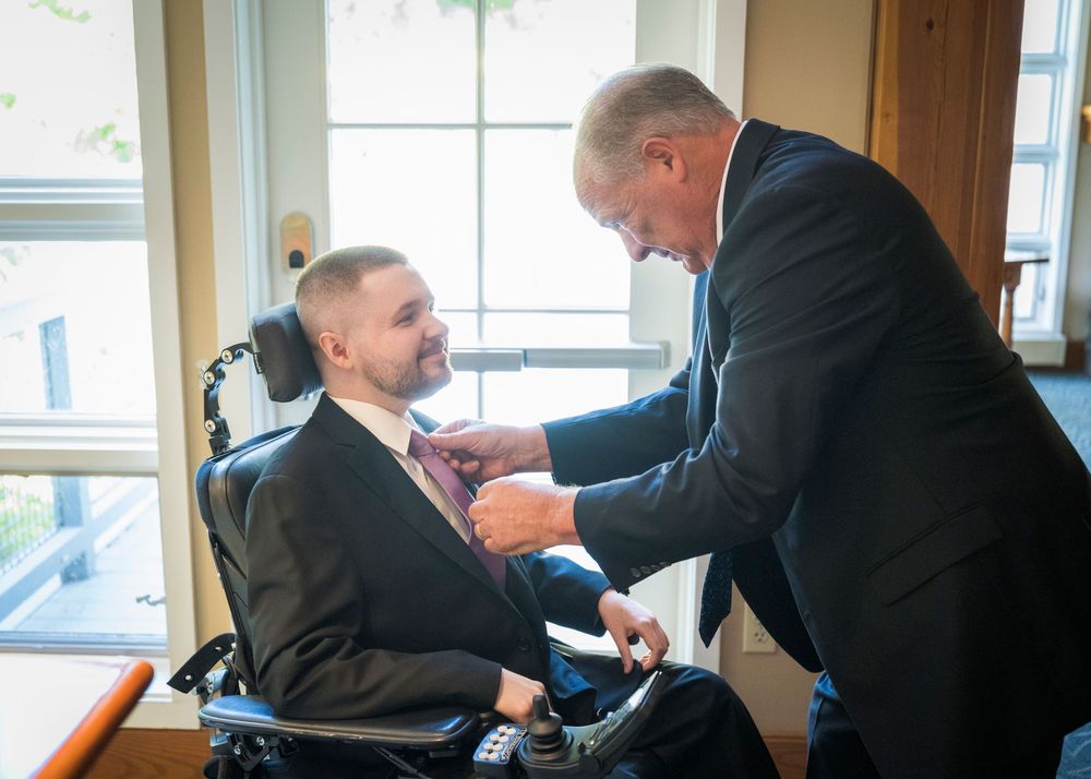 Man in wheelchair wearing suit gets tie adjusted by older man in dark suit near window.