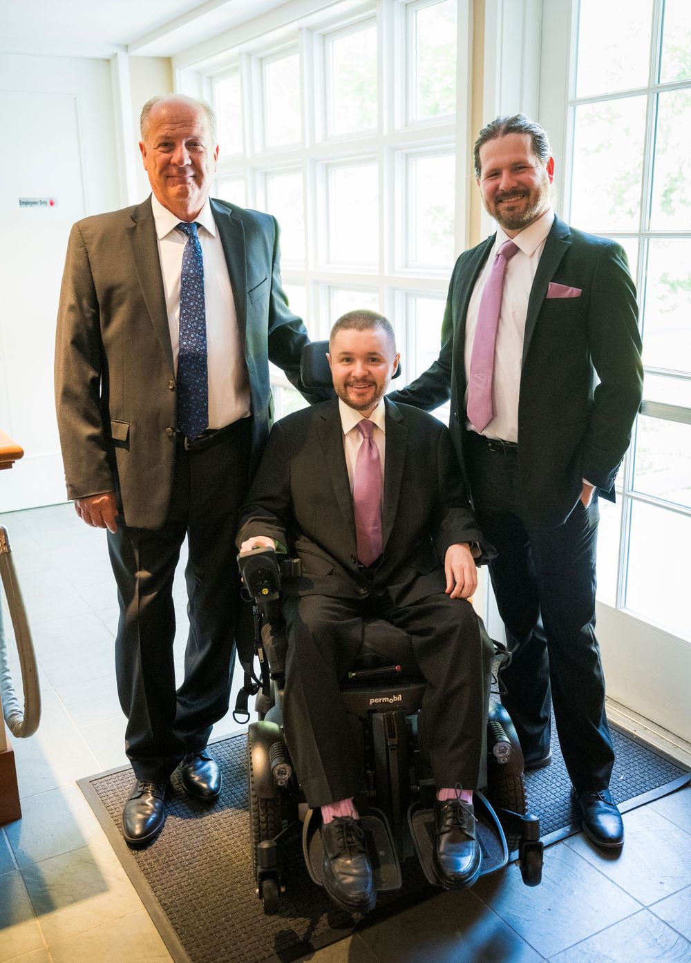 Three men in suits pose together, with one seated in a power wheelchair, smiling in a bright indoor setting.