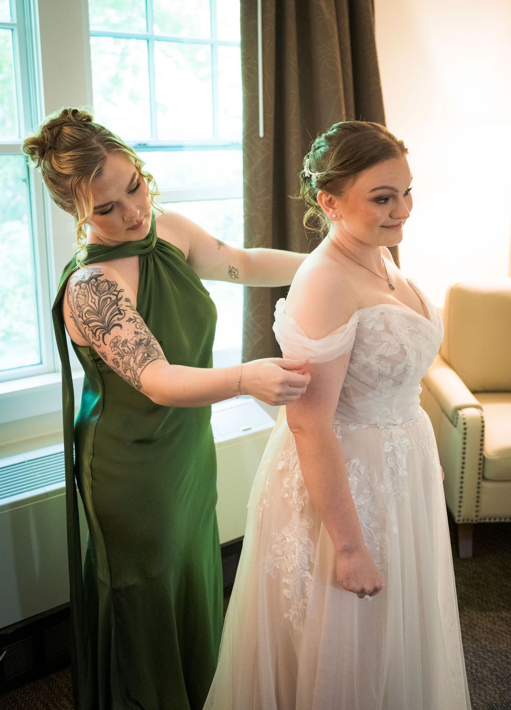 Bridesmaid in green halter dress with sleeve tattoo helps bride button her white lace off-shoulder wedding gown.