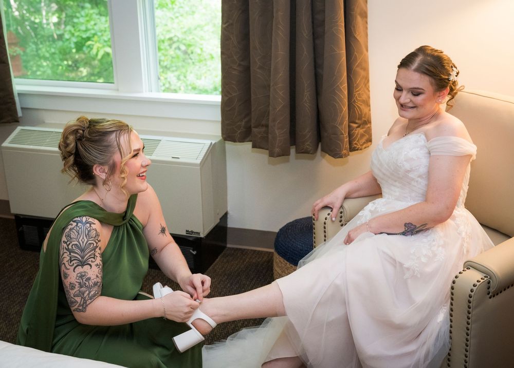 Bridesmaid in green dress helps bride with shoes in a hotel room before wedding ceremony.