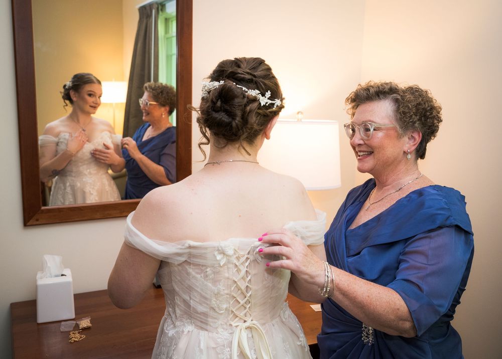 Bride in white wedding gown getting dressed with help from older woman in blue dress, reflected in mirror.