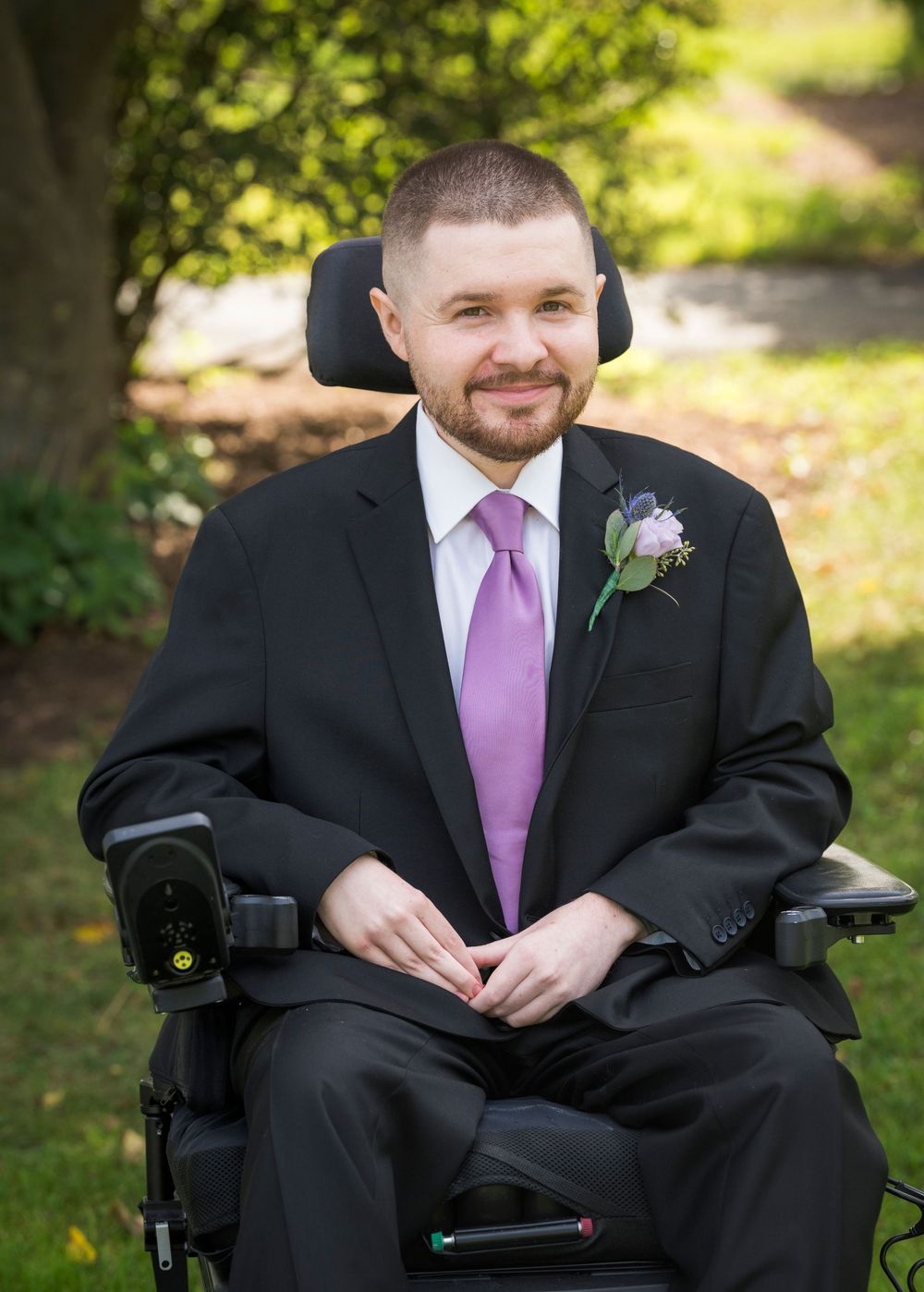 Smiling groom in black suit with purple tie and boutonniere seated in a power wheelchair outdoors.