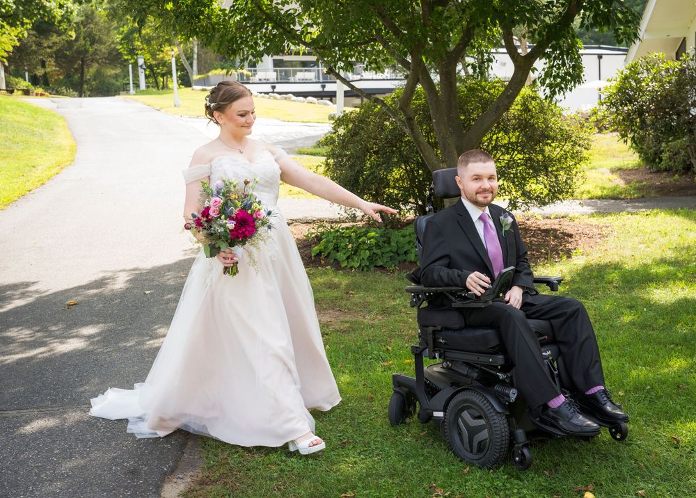 Bride in white gown holding bouquet walks beside groom in power wheelchair on tree-lined path at outdoor wedding.