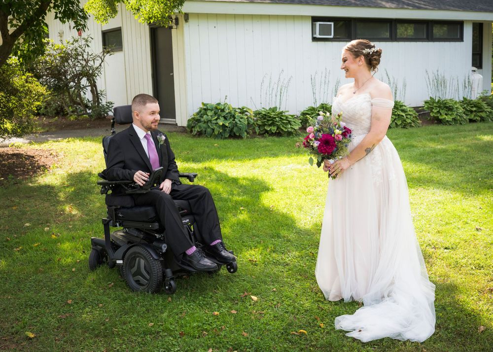 A groom in a wheelchair wearing a suit faces his bride in a white gown holding a bouquet on a green lawn.