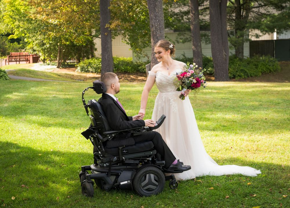 Bride in white gown holds bouquet beside groom in power wheelchair on green lawn during outdoor wedding ceremony.