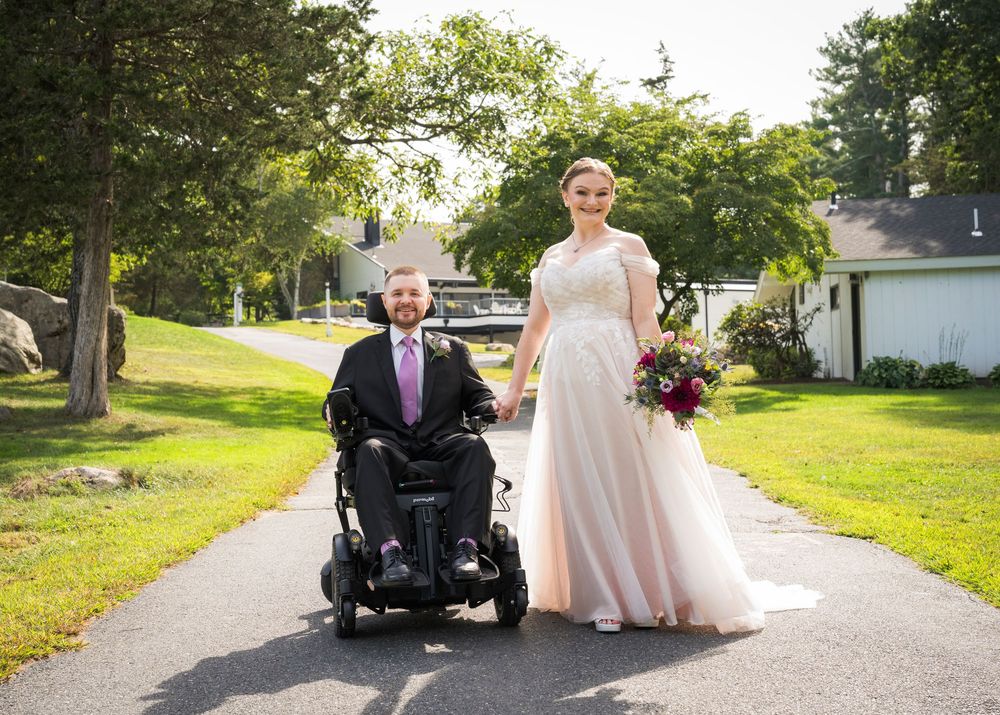 Bride in white gown stands beside groom in power wheelchair on sunny garden path on their wedding day.