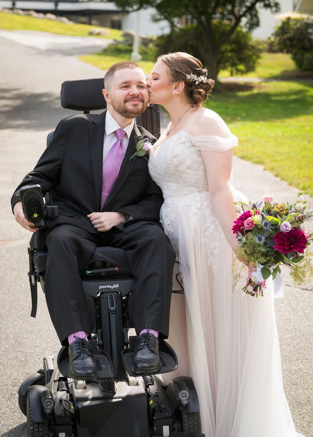 A groom in a wheelchair shares a tender kiss from his bride in a white gown holding a colorful bouquet outdoors.
